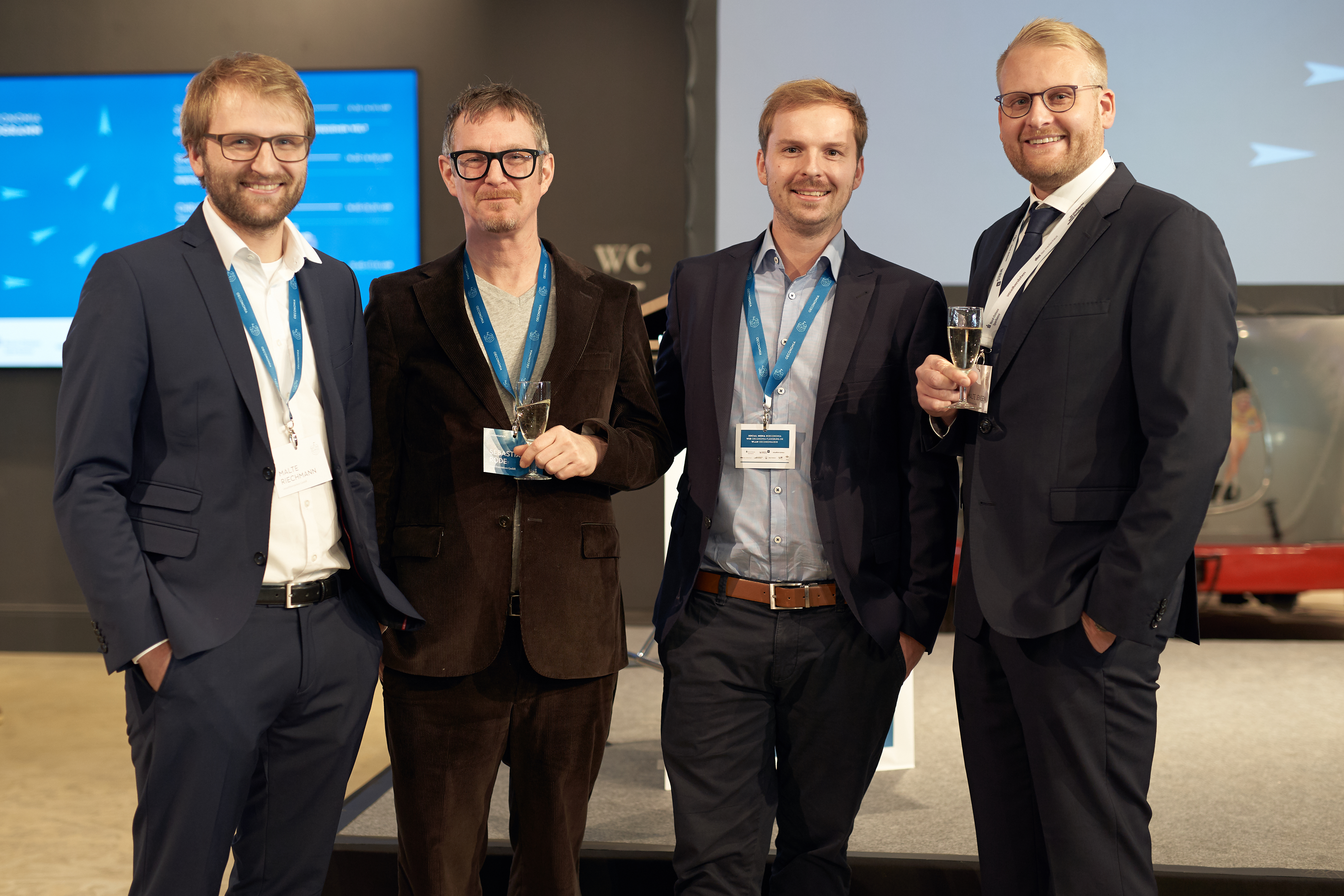 Die beiden Geschäftsführer und zwei Mitarbeiter von visuellverstehen posieren auf der Oeconomia 2018 mit Sektgläsern in der Hand für ein Foto.